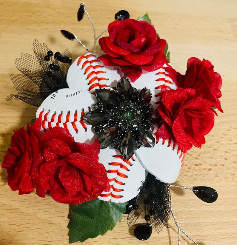 May include: A baseball-themed hair accessory with red fabric flowers, black netting, and a silver rhinestone embellishment. The baseballs have red stitching and the text "CENTER 9 IN."