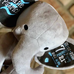 May include: Gray plush elephant stuffed animal with a black and blue embroidered tag on its ear. The tag reads "Zane Patrick" and includes the baby's birth information: "7 lbs 21 oz, 5:21 a.m., 01.01.2018".