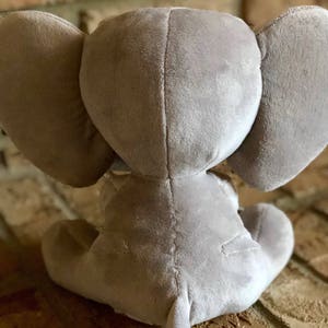 May include: A gray plush elephant toy sitting on a brick surface. The elephant has large ears and a long trunk.