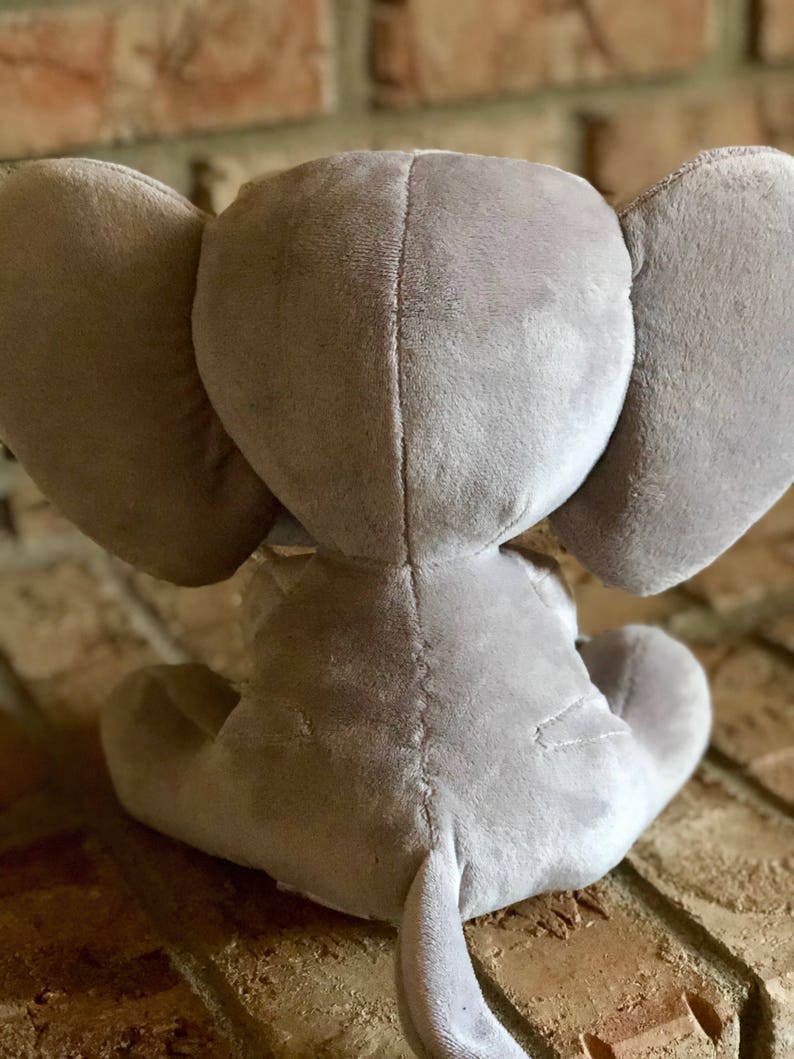 May include: A gray plush elephant toy sitting on a brick surface. The elephant has large ears and a long trunk.