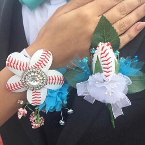 May include: Two baseball-themed boutonnieres, one with a white flower and a rhinestone center, the other with a white rose. Both have green leaves and blue accents.