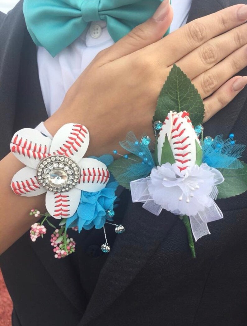 May include: Two baseball-themed boutonnieres, one with a white flower and a rhinestone center, the other with a white rose. Both have green leaves and blue accents.