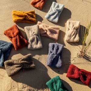 Puede incluir: Surtido de diademas de punto de colores, en varios colores, incluyendo azul, verde, rojo, marr&oacute;n y beige. Las diademas est&aacute;n dispuestas sobre una superficie arenosa.