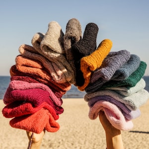 Pode incluir: Uma pilha arco-íris de chapéus de tricô em várias cores, incluindo rosa, vermelho, laranja, amarelo, verde, azul, roxo e cinza. Os chapéus estão dispostos em forma de arco e são segurados por duas mãos.