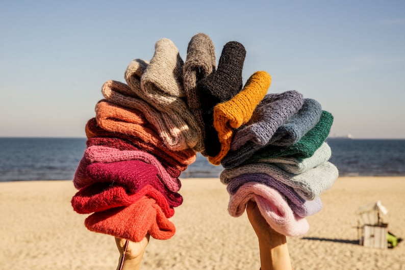 Puede incluir: Una pila arco&iacute;ris de gorros de punto en varios colores, incluyendo rosa, rojo, naranja, amarillo, verde, azul, morado y gris. Los gorros est&aacute;n dispuestos en forma de arco y son sostenidos por dos manos.