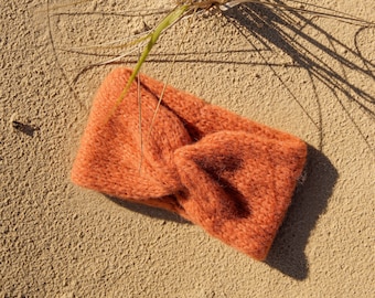 Puede incluir: Una diadema de punto naranja suave con un diseño de nudo retorcido. La diadema está sobre una superficie arenosa.