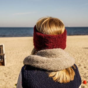 Puede incluir: Una mujer con una diadema de punto roja y una bufanda gris est&aacute; de pie en una playa de arena mirando al mar.