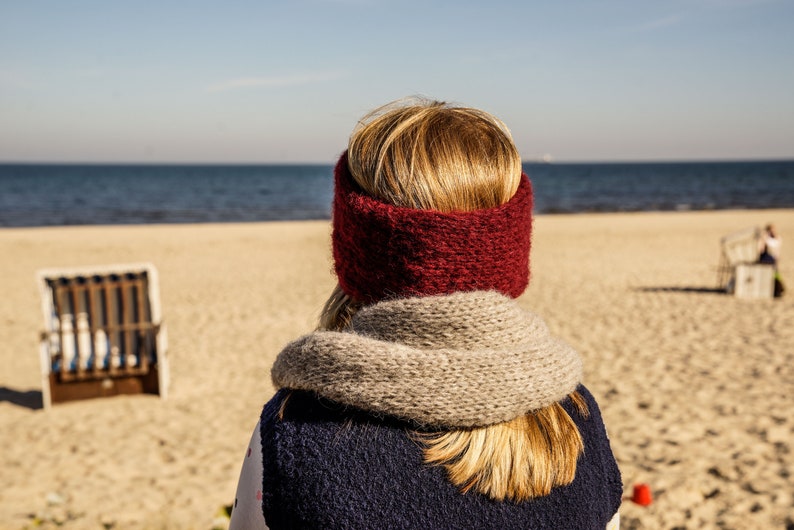Puede incluir: Una mujer con una diadema de punto roja y una bufanda gris est&aacute; de pie en una playa de arena mirando al mar.