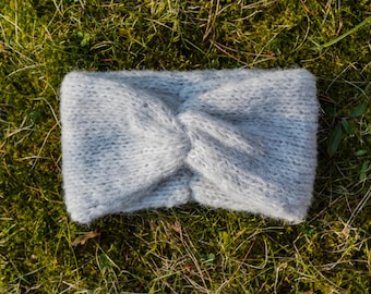 Op de afbeelding: Een grijs gebreid hoofdband met een geknoopt ontwerp. Het hoofdband ligt op een bed van groen mos.