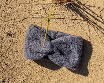 Op de afbeelding: Een donkergrijze gebreide hoofdband met een geknoopt ontwerp. De hoofdband ligt op een zanderig oppervlak.