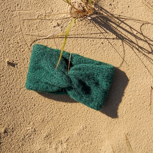 Op de afbeelding: Een donker bosgroene gebreide hoofdband met een centraal gedraaid knoopdetail. Het accessoire is gemaakt van zacht, pluizig garen met een zichtbaar gestructureerd breipatroon, wat zorgt voor een dikke en warme uitstraling van dit hoofddeksel.