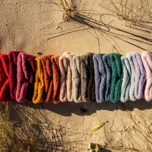 Puede incluir: Una fila de 12 diademas de punto de colores en varios tonos de rojo, rosa, naranja, beige, gris, azul, verde y morado. Las diademas est&aacute;n dispuestas en l&iacute;nea sobre una superficie arenosa.