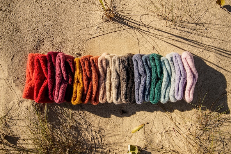 Puede incluir: Una fila de 12 diademas de punto de colores en varios tonos de rojo, rosa, naranja, beige, gris, azul, verde y morado. Las diademas est&aacute;n dispuestas en l&iacute;nea sobre una superficie arenosa.