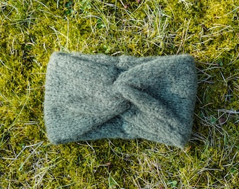 Op de afbeelding: Een groene gebreide hoofdband met een gedraaid knoopontwerp. De hoofdband ligt op een bed van groen mos.