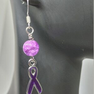May include: A single dangle earring featuring a purple ribbon charm and a textured purple bead. The earring has a silver-colored hook and connecting elements. The ribbon charm is outlined in silver.
