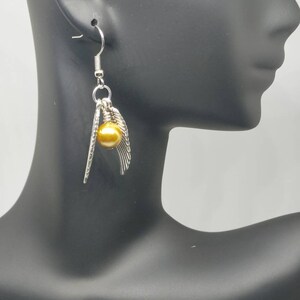 May include: Silver-toned dangle earrings featuring a golden sphere and feather-like accents. The earrings are suspended from a simple hook and are displayed on a black mannequin head. The jewelry has a delicate and elegant design.