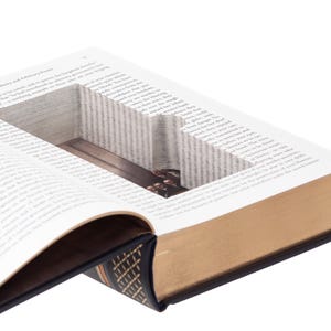 May include: A hardcover book with a rectangular hollow compartment cut into the center of its pages. The book features a dark blue cover with gold-colored spine detailing and gold-edged pages. The interior cavity reveals a smooth, dark surface.