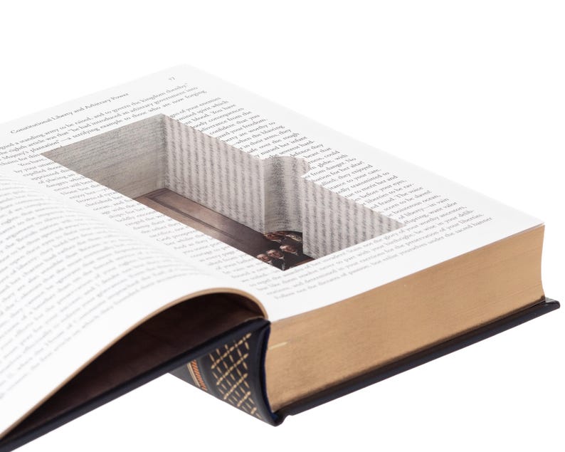 May include: A hardcover book with a rectangular hollow compartment cut into the center of its pages. The book features a dark blue cover with gold-colored spine detailing and gold-edged pages. The interior cavity reveals a smooth, dark surface.