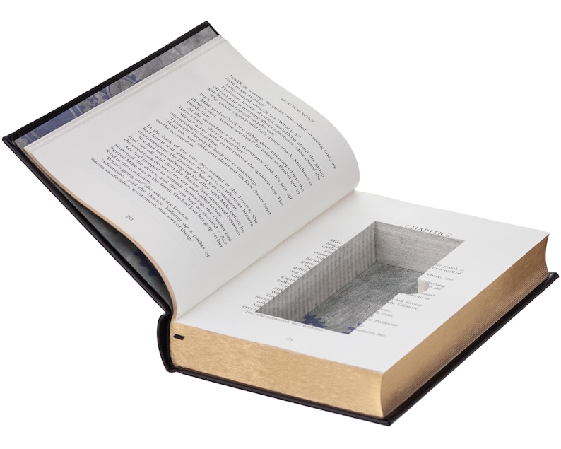 May include: A hardcover book with a rectangular hollow compartment carved into the center of its pages. The book is open to Chapter 2, revealing white paper pages with black printed text and gold-toned edges on the outer side of the pages.