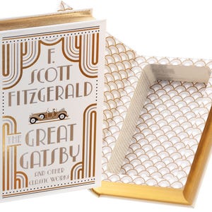 Puede incluir: Una caja fuerte de libro blanca con un diseño dorado y el título "The Great Gatsby and Other Classic Works" de F. Scott Fitzgerald. El libro se abre para revelar un compartimento oculto.