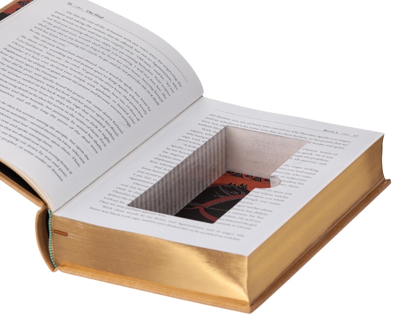 May include: An open hardcover book with gold-gilded page edges features a rectangular hollow compartment cut into the center of the pages. The interior of the hidden cavity reveals a dark red and black patterned lining with a graphic design.
