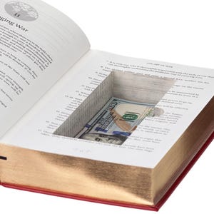 May include: A hollowed-out hardcover book titled The Art of War with a red cover and gold-gilded page edges. A rectangular compartment is cut into the interior pages, revealing a folded one hundred dollar bill tucked inside the hidden space.