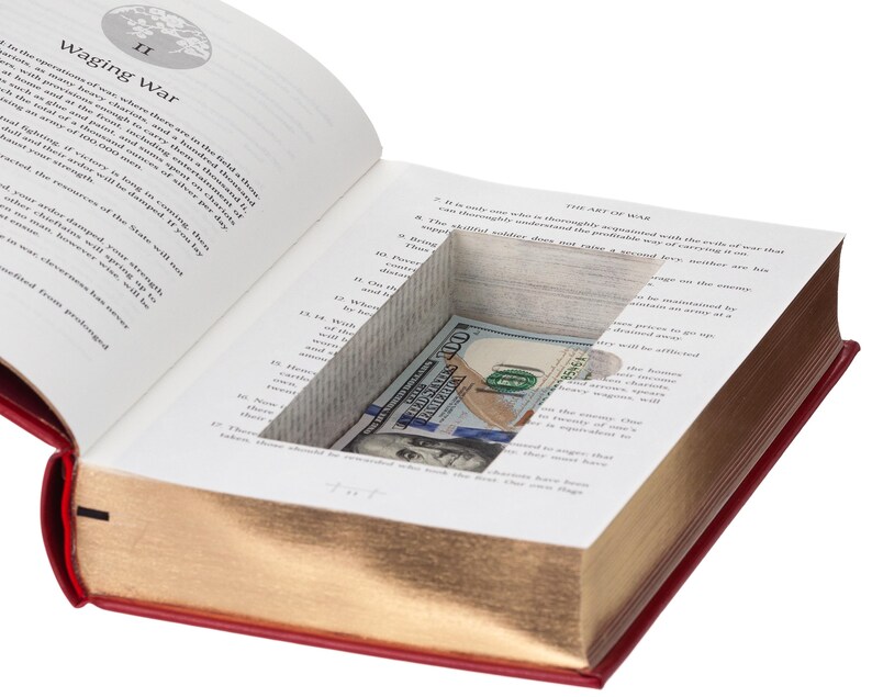May include: A hollowed-out hardcover book titled The Art of War with a red cover and gold-gilded page edges. A rectangular compartment is cut into the interior pages, revealing a folded one hundred dollar bill tucked inside the hidden space.