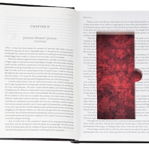 May include: An open hardcover book titled Dracula, featuring a rectangular cutout in the right page that reveals a deep red interior lining with a dark, ornate damask pattern. The left page displays printed text under the heading Chapter II.