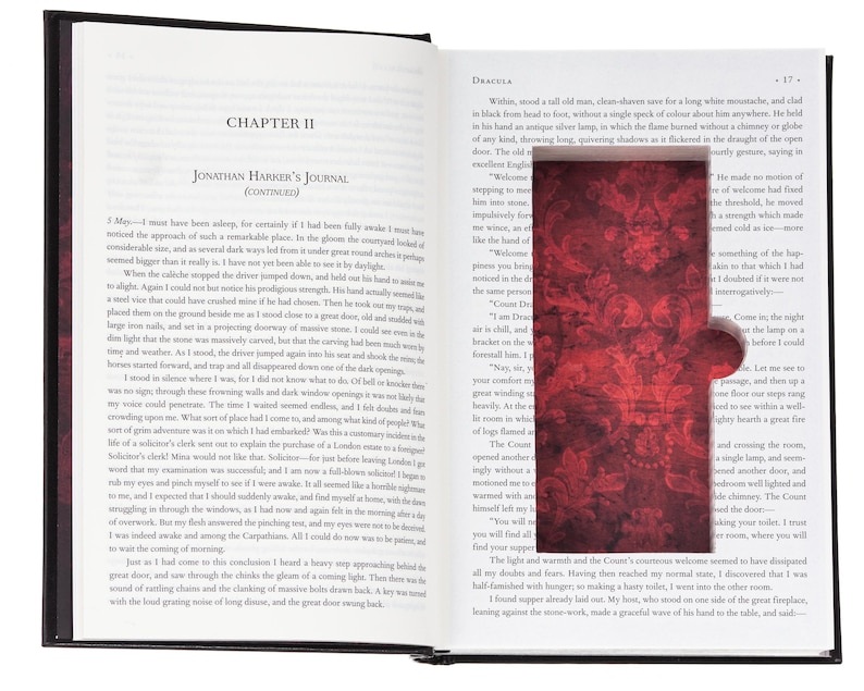 May include: An open hardcover book titled Dracula, featuring a rectangular cutout in the right page that reveals a deep red interior lining with a dark, ornate damask pattern. The left page displays printed text under the heading Chapter II.