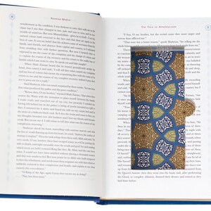 May include: An open hardcover book with printed text on white pages. A rectangular cutout in the right page reveals a bookmark featuring a repeating geometric pattern in shades of blue, gold, and white with a small circular tab on the side.