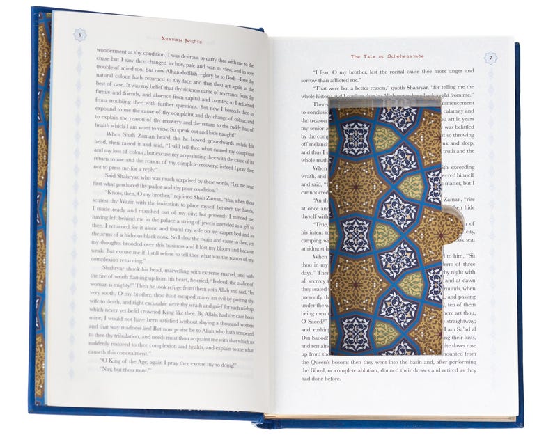 May include: An open hardcover book with printed text on white pages. A rectangular cutout in the right page reveals a bookmark featuring a repeating geometric pattern in shades of blue, gold, and white with a small circular tab on the side.