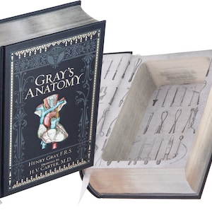 Puede incluir: Una caja negra con forma de libro con el título "Gray's Anatomy" en la portada. La portada presenta una ilustración de un corazón humano y el texto "Henry Gray, F.R.S. Dibujos de H.V. Carter, M.D.". La caja está abierta para revelar un compartimento oculto.