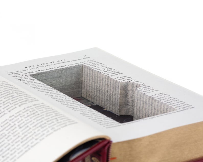 May include: A hardcover book with a rectangular hollow compartment carved into the center of its pages. The visible text at the top of the page reads The Ages of Man. The book features a deep red spine and gold-toned page edges.