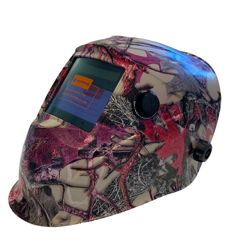 May include: A pink and grey camouflage welding helmet with a digital display and a warning label that reads "Please Remove This Welding Filter Prior to Use".