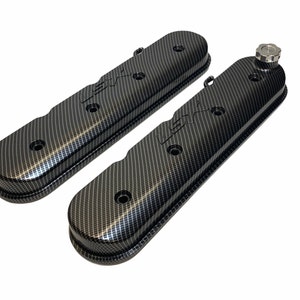 May include: A pair of black carbon fiber valve covers with the text "LSX" embossed on each cover. The covers have a textured finish and are designed for use on a car engine.