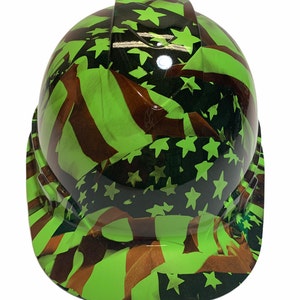 Könnte beinhalten: Ein grüner Schutzhelm mit einem Tarnmuster der amerikanischen Flagge. Die Flagge ist in Grün-, Schwarz- und Brauntönen gehalten.
