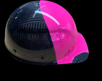 Può includere: Un casco di sicurezza con un design diviso. Una metà è rosa acceso lucido, l'altra è nera con un motivo in fibra di carbonio. La visiera è anch'essa rosa con un lato inferiore strutturato.