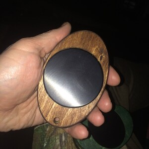 May include: A wooden oval-shaped ornament with a round, black, metal disc in the center. The ornament has a small hole at the top.