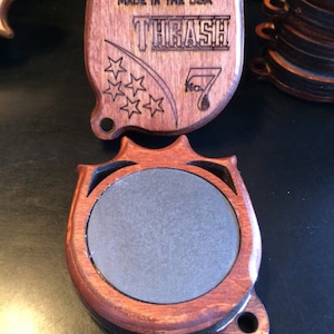 May include: A wooden, brown, and black, engraved, circular,  medallion with the text "BAGREBUCK.COM MADE IN THE USA THRASH No. 7" and five stars. The medallion has a gray, circular,  felt-like insert.