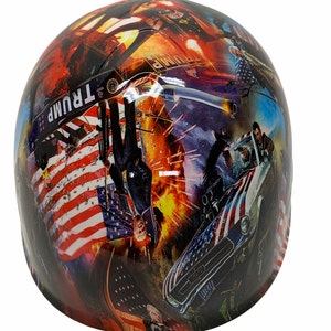 May include: A hard hat with a colorful collage design featuring an American flag, a car, and a man in a suit. The design also includes the word "TRUMP" and the text "LIBERTY".