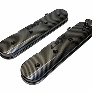 May include: Two carbon fiber-style engine valve covers. The covers are black with a woven pattern and the letters "LSX" in black. The covers have several bolt holes and a raised edge.