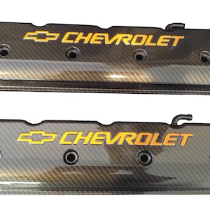 Könnte beinhalten: Ein Paar schwarze Ventildeckel aus Kohlefaser mit goldenem Chevrolet-Logo. Die Deckel haben eine strukturierte Oberfläche und sind für einen Automotor konzipiert.
