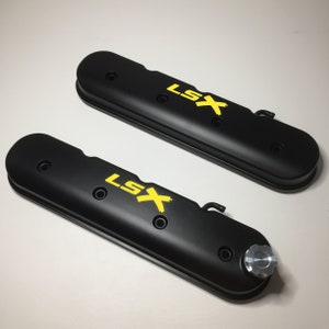 Könnte beinhalten: Zwei schwarze Ventildeckel mit gelber "LSX" Beschriftung. Die Ventildeckel haben eine strukturierte Oberfläche und sind für einen Automotor konzipiert.