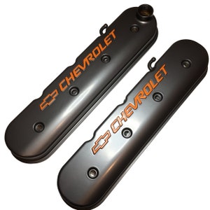 Könnte beinhalten: Ein Paar schwarze Ventildeckel mit orangefarbener "Chevrolet"-Beschriftung. Die Deckel haben eine glatte, metallische Oberfläche.