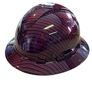 Puede incluir: Un casco de seguridad rojo y negro con un patrón de fibra de carbono. El casco tiene un ala ancha y una correa negra.