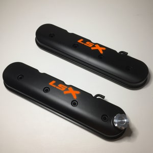 May include: Two black engine valve covers with orange "LSX" lettering. The valve covers have a textured finish and are designed for a car engine.