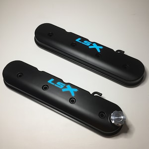 Könnte beinhalten: Zwei schwarze Ventildeckel mit einem blauen "LSX"-Logo. Die Ventildeckel haben eine strukturierte Oberfläche und sind für einen Automotor konzipiert.