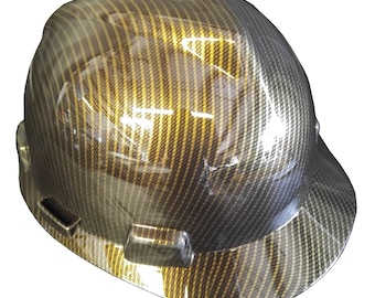 May include: A hard hat with a black and gold carbon fiber pattern. The hard hat has a brim and a chin strap.