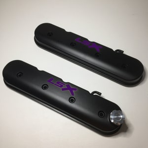 May include: Two black valve covers with purple "LSX" lettering. The valve covers have a textured finish and are designed for a car engine.