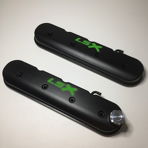 May include: Two black valve covers with green "LSX" logos. The valve covers have multiple holes and a silver knob on one of the covers.